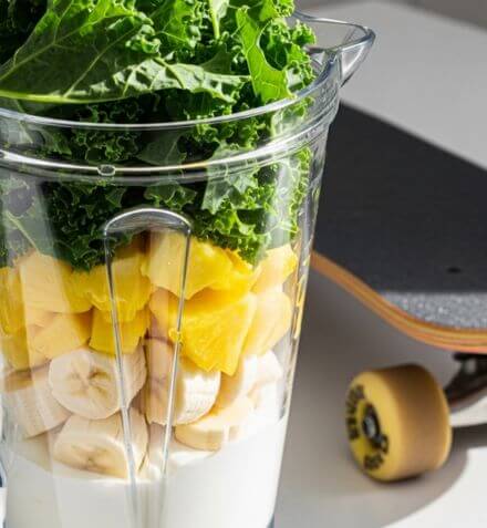 Blender filled with milk, banana, pineapple and kale beside a skateboard, ready to make a green smoothie.