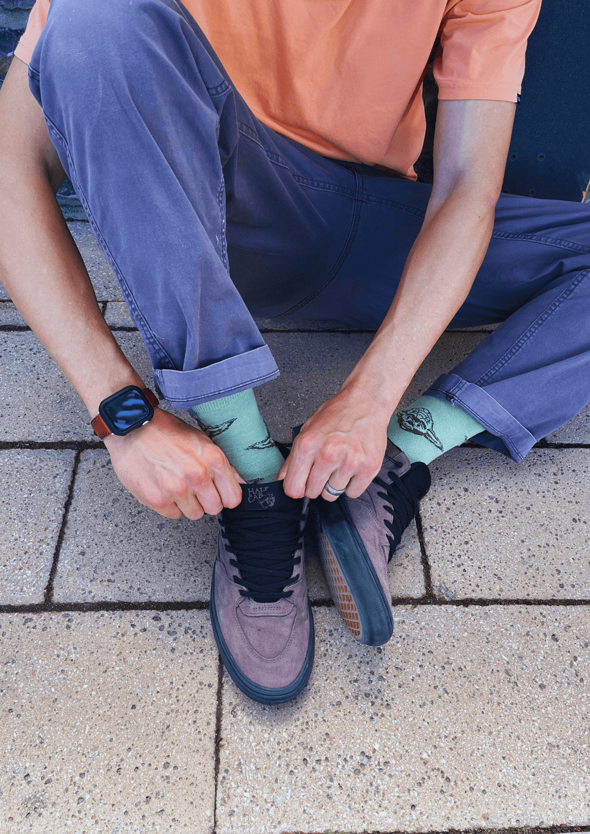 Skateboarder sitting on pavement and tying black skate shoes while wearing blue trousers, green socks and an orange T-shirt.